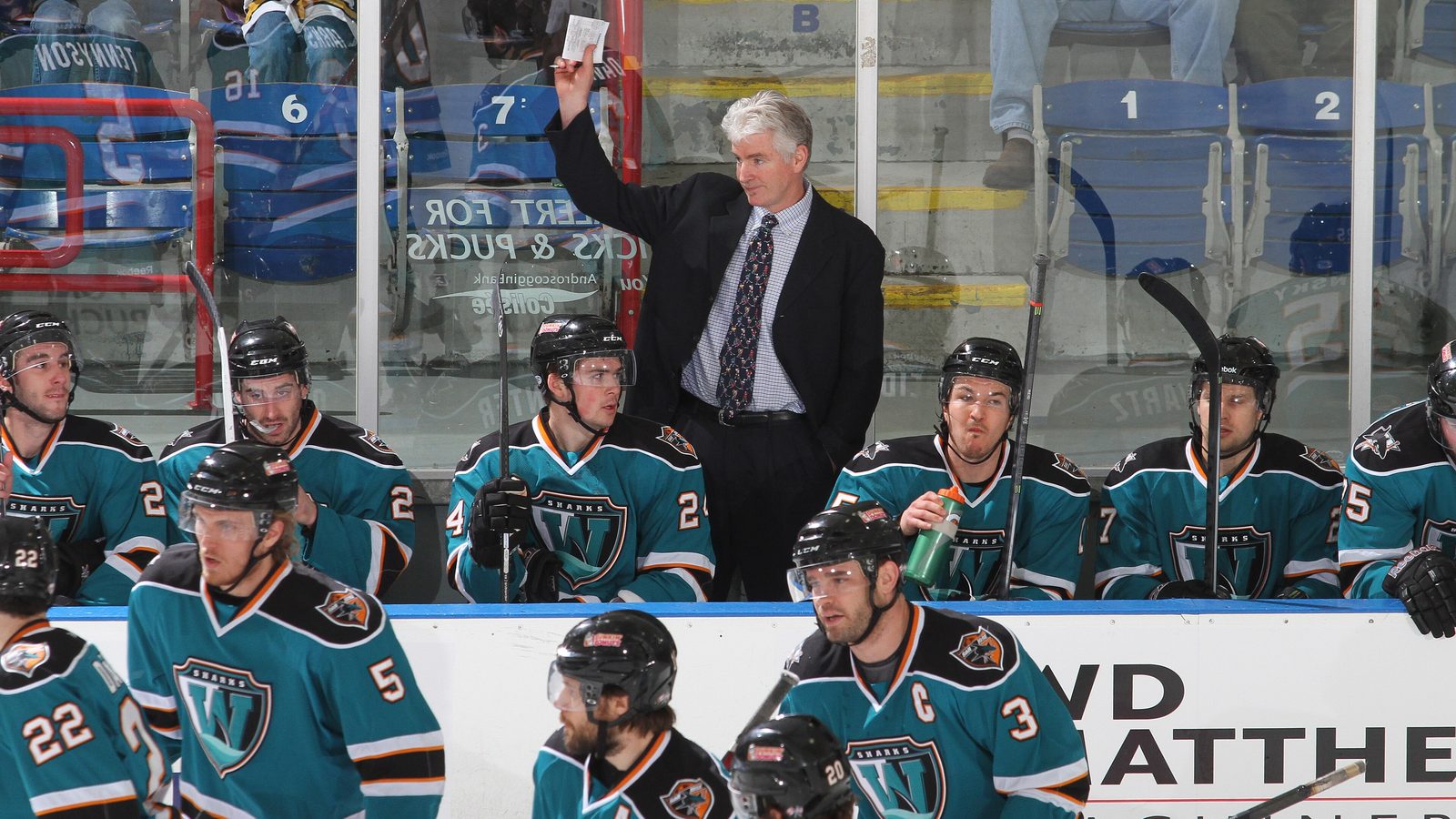 A coach in a suit stands behind a bench of seated players in teal Worcester Sharks jerseys