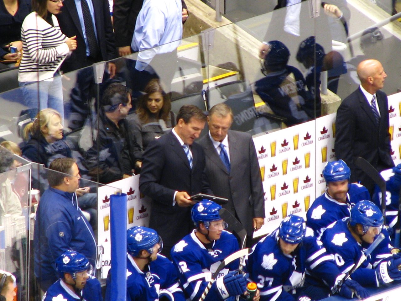 Two coaches in suits look at a tablet behind the Toronto Maple Leafs bench during a game