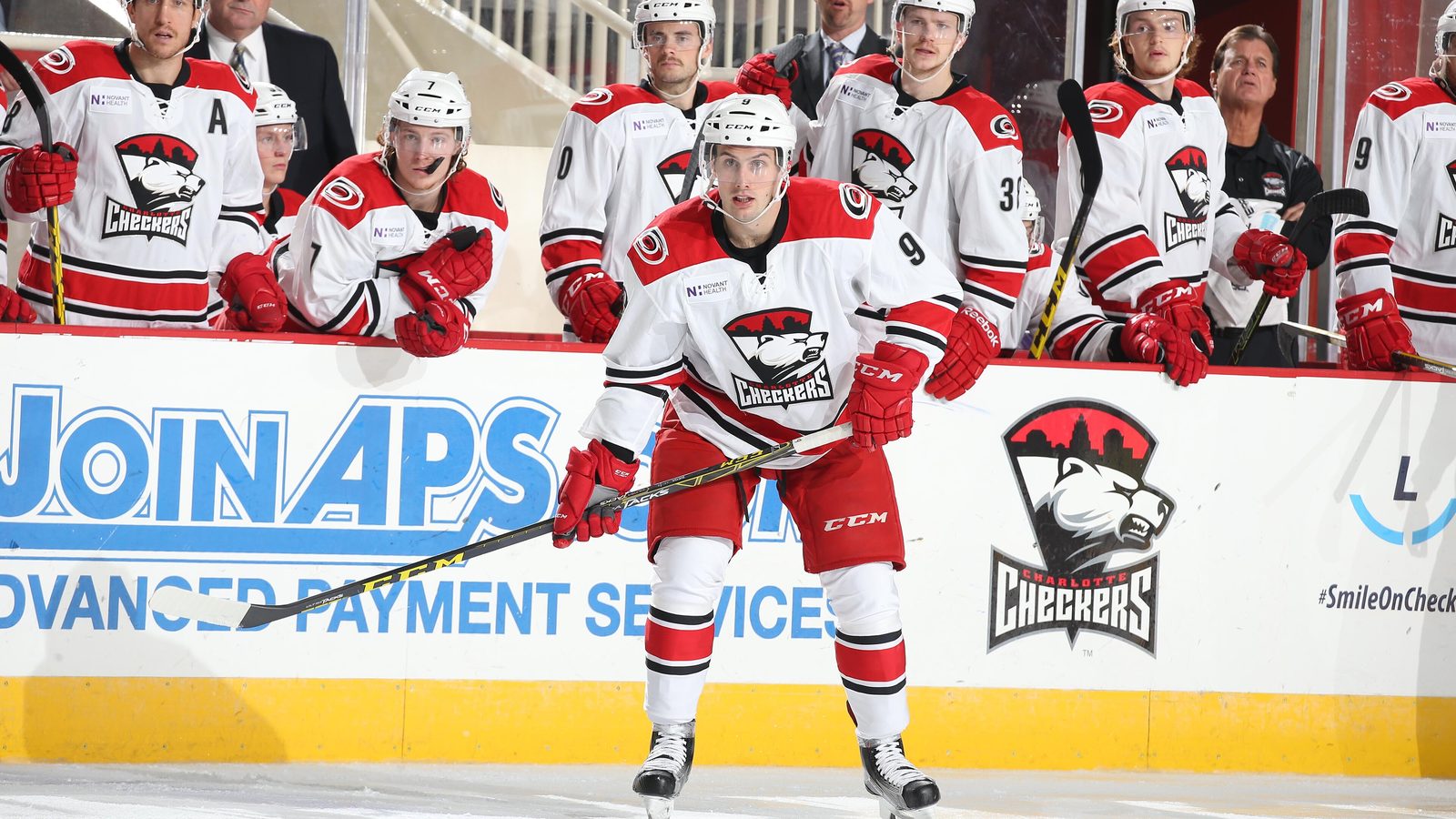 A Charlotte Checkers player in a white jersey skates in front of his team's bench, with staff in suits standing behind the players