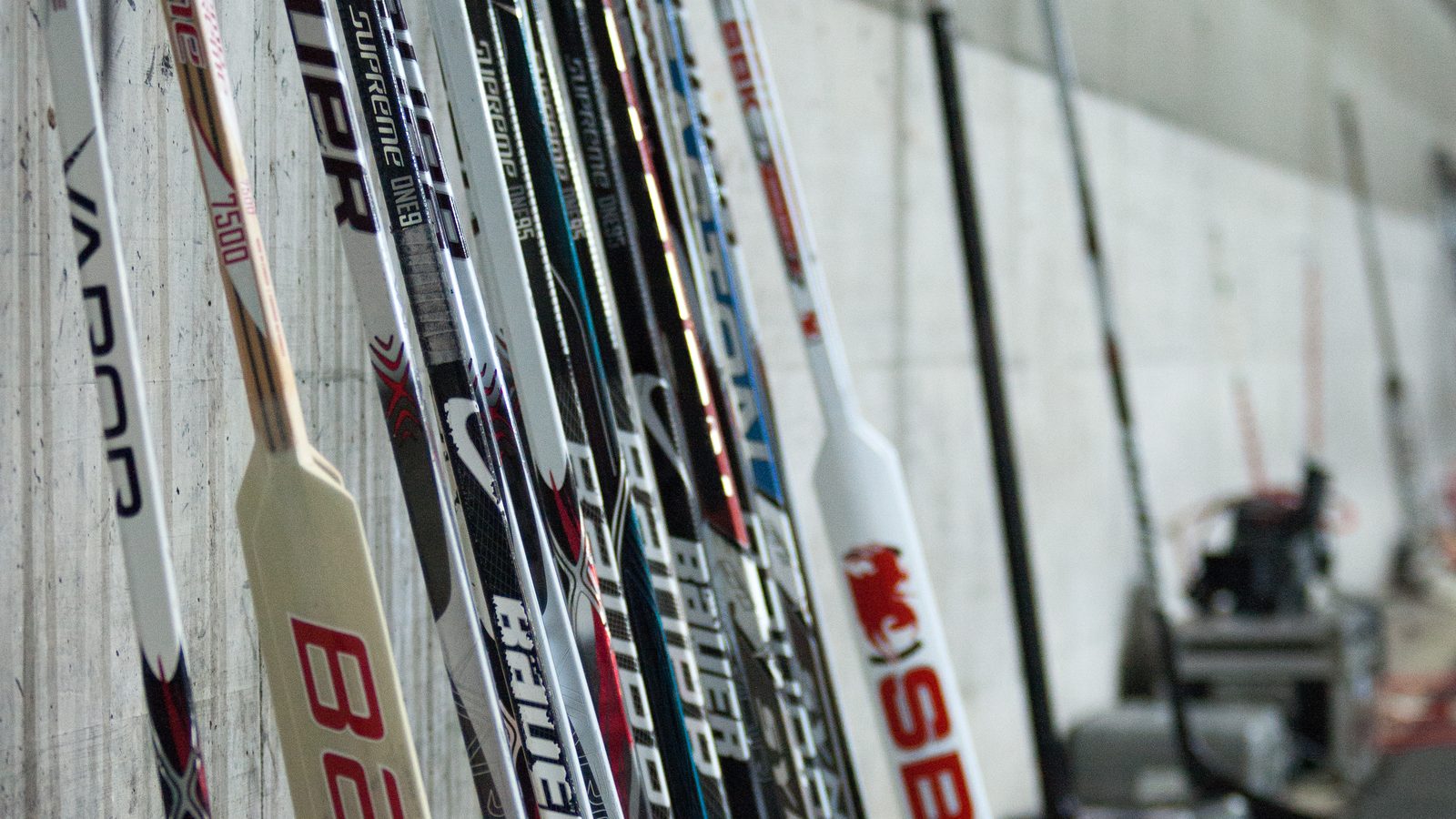 A row of hockey sticks leaning against a concrete wall in an arena corridor